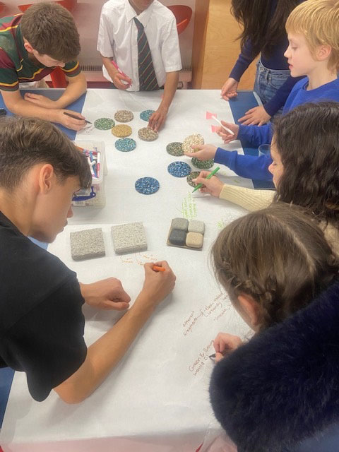 Workshop participants are pictured standing around a table, examining objects on a table and writing notes 